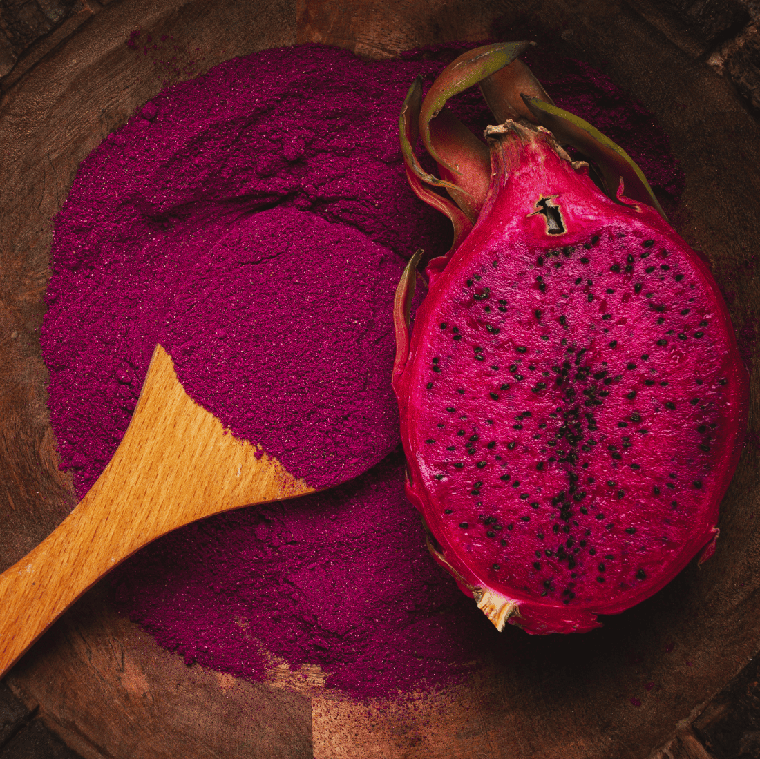 Bright pink dragon fruit powder with a wooden spoon and a halved red dragon fruit on a wooden surface.