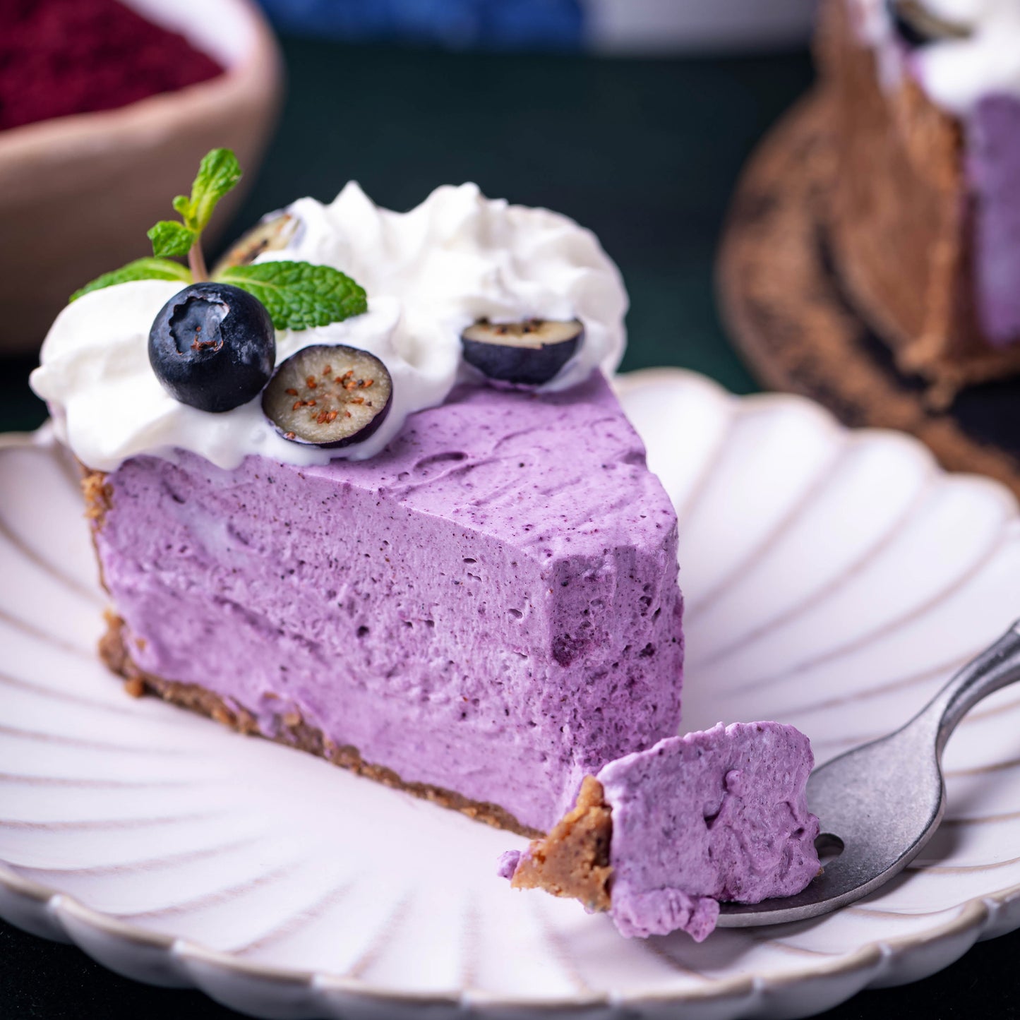 Slice of blueberry cheesecake made with Ancient Choice Wild Blueberry Powder, topped with whipped cream, fresh blueberries, and a mint leaf on a white plate.