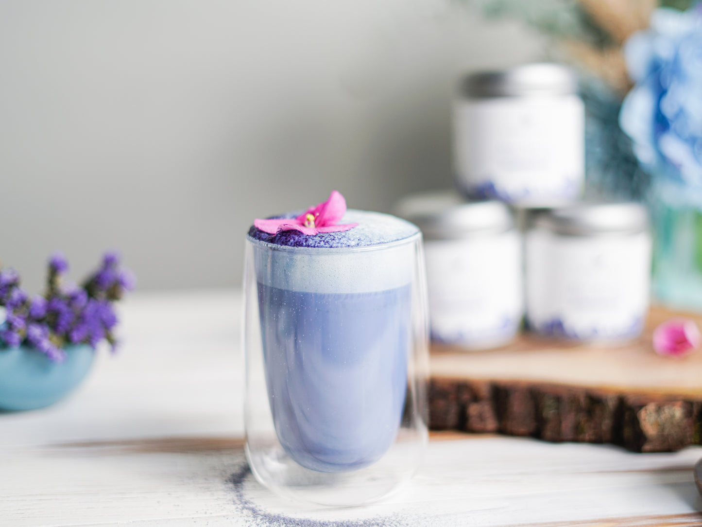 Blue matcha latte made with Ancient Choice Blue Matcha, topped with foam and a pink flower garnish in a clear double-wall glass.