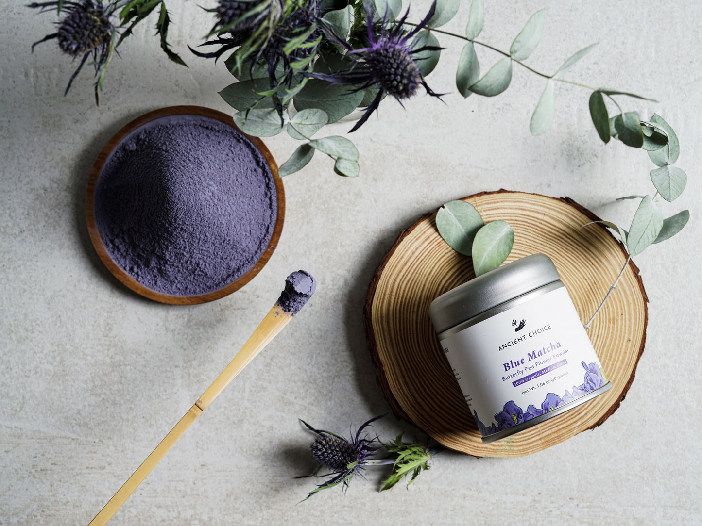 Ancient Choice Blue Matcha powder displayed in a wooden bowl and tin canister, with a bamboo spoon and botanical accents.