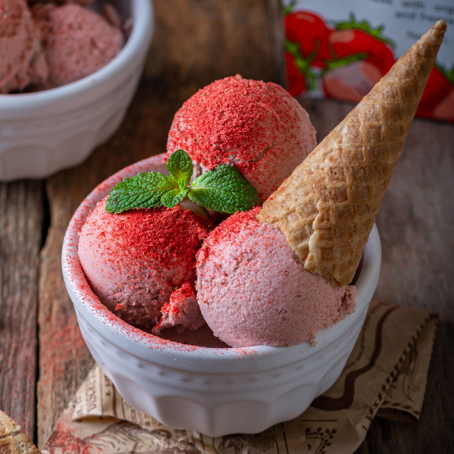 Strawberry ice cream made with Ancient Choice Strawberry Powder, served in a white bowl with a waffle cone, mint garnish, and powder dusting on top.