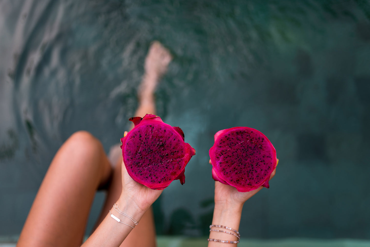 Hands holding two halves of a bright pink dragon fruit over water.