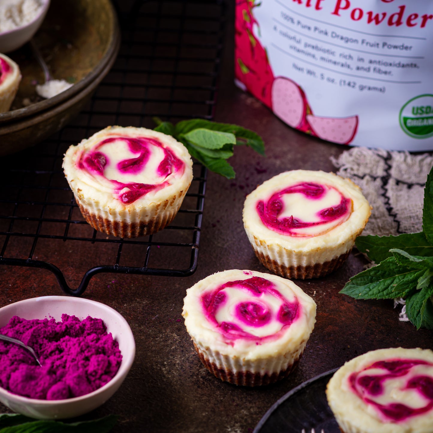 Mini cheesecakes made with Ancient Choice Dragon Fruit Powder, displayed on a cooling rack and rustic table with mint leaves and a bowl of dragon fruit powder nearby.