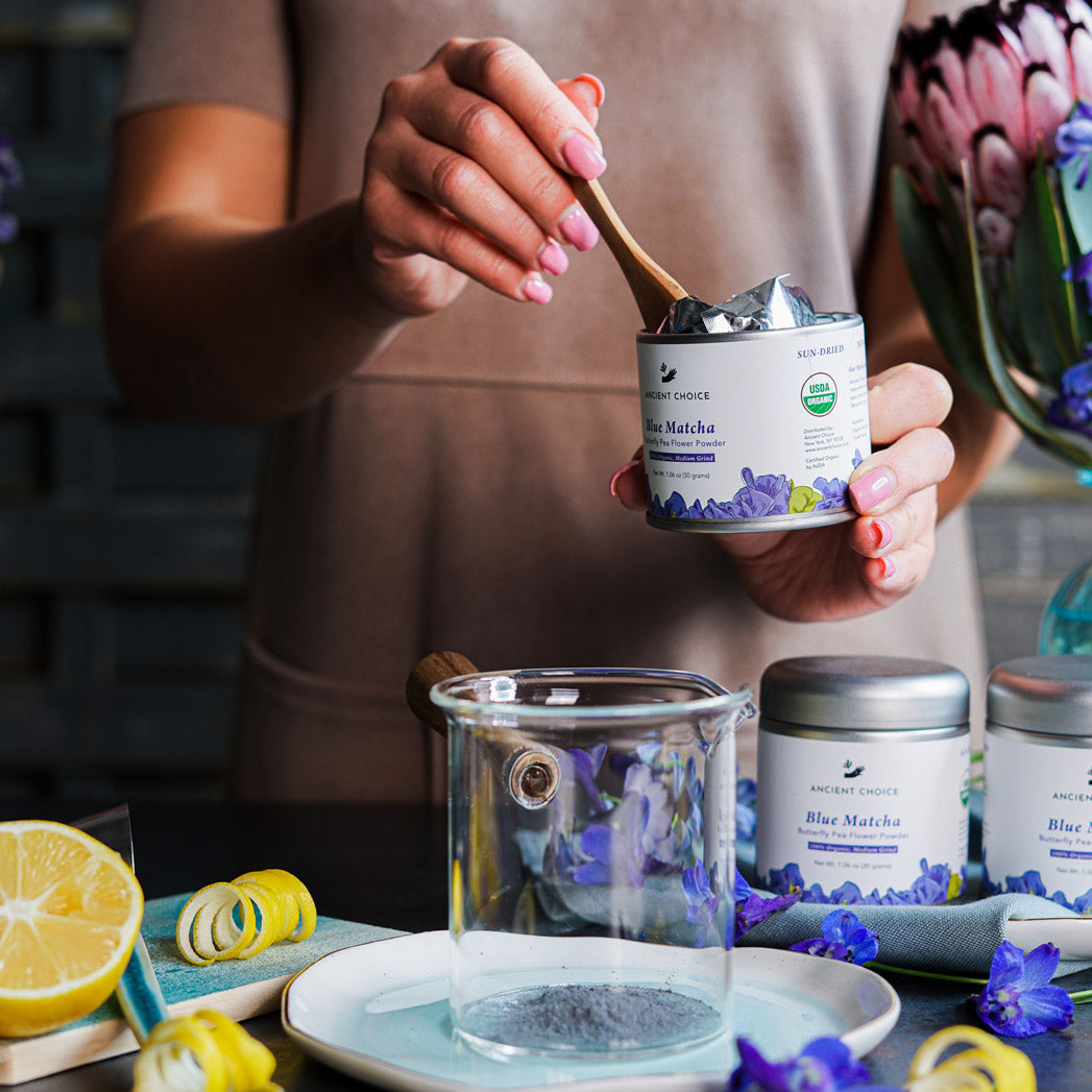 Scooping Ancient Choice Blue Matcha into a glass, surrounded by lemons and fresh butterfly pea flowers.